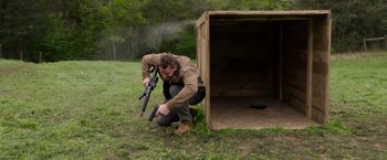 Movie still from “The Hunt” (2020), directed by Craig Zobel – A man kneeling in front of a wooden box holding a rifle; Wide shot, Low angle