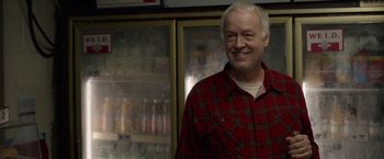 Movie still from “The Hunt” (2020), directed by Craig Zobel – An older man wearing a plaid shirt standing in front of a refrigerator; Medium shot, Over the shoulder angle