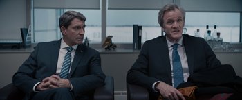 Movie still from “The Ice Road” (2021), directed by Jonathan Hensleigh – Two men in suits sitting next to each other in front of a window; Medium shot, Over the shoulder angle
