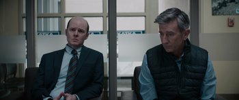 Movie still from “The Ice Road” (2021), directed by Jonathan Hensleigh – Two men in suits sitting next to each other in a room; Medium shot, Over the shoulder angle