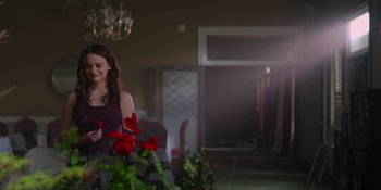 Movie still from “The In Between” (2022), directed by Arie Posin – A beautiful young lady standing next to flowers in a room; Medium shot, Over the shoulder angle