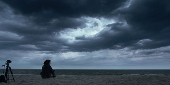 Movie still from “The In Between” (2022), directed by Arie Posin – A person sitting on a beach under a cloudy sky; Extreme Wide shot, Low angle