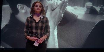 Movie still from “The In Between” (2022), directed by Arie Posin – A woman standing in front of a large picture of hands; Medium shot, Over the shoulder angle