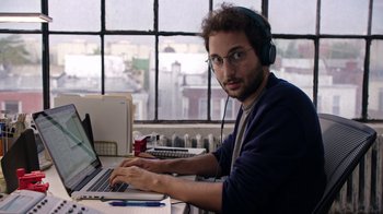 Movie still from “The Intern” (2015), directed by Nancy Meyers – A man wearing headphones and sitting at a desk with a computer; Medium shot, Over the shoulder angle