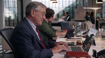 Movie still from “The Intern” (2015), directed by Nancy Meyers – A man sitting at a table using a laptop computer; Medium shot, Over the shoulder angle