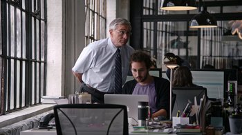 Movie still from “The Intern” (2015), directed by Nancy Meyers – A man standing next to another man in front of a computer; Medium shot, Over the shoulder angle