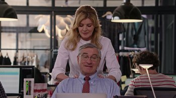 Movie still from “The Intern” (2015), directed by Nancy Meyers – A woman standing behind a man sitting on his shoulders; Medium shot, Over the shoulder angle