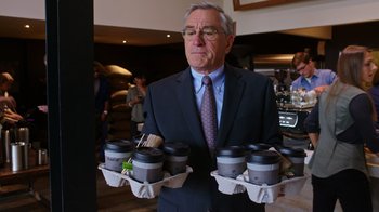 Movie still from “The Intern” (2015), directed by Nancy Meyers – An older man in a business suit holding a tray of coffee cups; Medium shot, Over the shoulder angle