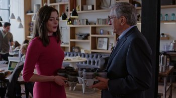 Movie still from “The Intern” (2015), directed by Nancy Meyers – A woman in a red dress and a man in a gray suit holding bowls; Medium shot, Over the shoulder angle
