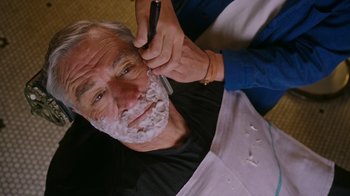 Movie still from “The Intern” (2015), directed by Nancy Meyers – An older man getting his beard shaved by a barber; Close Up shot, Over the shoulder angle