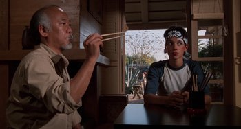 Movie still from “The Karate Kid Part II” (1986), directed by John G. Avildsen – Two people sitting at a table with chopsticks in their mouth; Medium shot, Over the shoulder angle