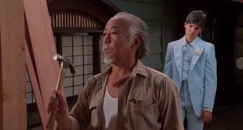 Movie still from “The Karate Kid Part II” (1986), directed by John G. Avildsen – An older man holding a hammer while standing in front of another man in a suit; Medium shot, Low angle