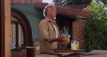 Movie still from “The Karate Kid Part II” (1986), directed by John G. Avildsen – An older man standing at a table holding a bowl of food; Medium shot, Low angle