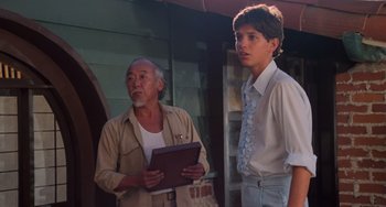 Movie still from “The Karate Kid Part II” (1986), directed by John G. Avildsen – A man standing next to a young man holding a tablet; Medium shot, Over the shoulder angle