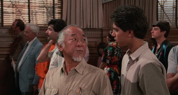 Movie still from “The Karate Kid Part II” (1986), directed by John G. Avildsen – A man with a white beard standing next to another man; Medium shot, Over the shoulder angle
