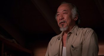 Movie still from “The Karate Kid Part II” (1986), directed by John G. Avildsen – An older man with a beard is smiling for the camera; Medium shot, Low angle