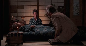 Movie still from “The Karate Kid Part II” (1986), directed by John G. Avildsen – An older man and a younger woman sitting on a bed; Wide shot, High angle