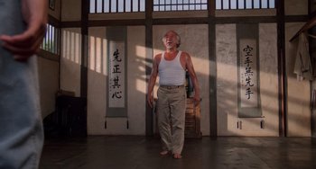 Movie still from “The Karate Kid Part II” (1986), directed by John G. Avildsen – An older man standing in a room with a white tank top on; Wide shot, Low angle