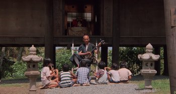 Movie still from “The Karate Kid Part II” (1986), directed by John G. Avildsen – A group of people sitting on the ground with a man playing a guitar; Wide shot, High angle