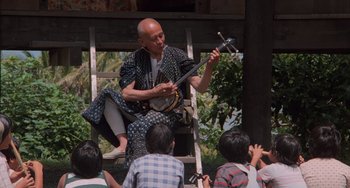 Movie still from “The Karate Kid Part II” (1986), directed by John G. Avildsen – An older man playing an instrument while sitting on a ladder; Medium shot, Low angle