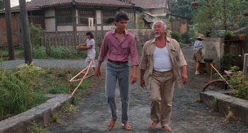 Movie still from “The Karate Kid Part II” (1986), directed by John G. Avildsen – A man standing next to another man on a dirt road; Wide shot, Low angle