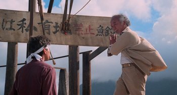 Movie still from “The Karate Kid Part II” (1986), directed by John G. Avildsen – A man standing next to another man in front of a wooden sign; Wide shot, Low angle