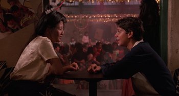Movie still from “The Karate Kid Part II” (1986), directed by John G. Avildsen – Two people sitting at a table in front of a crowd of onlookers; Medium shot, Over the shoulder angle