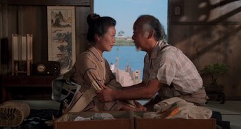 Movie still from “The Karate Kid Part II” (1986), directed by John G. Avildsen – An older man and a younger woman sitting on a bed in front of a window; Medium shot, Low angle