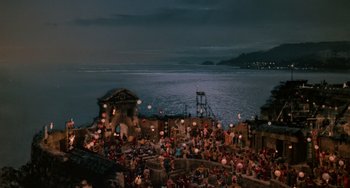 Movie still from “The Karate Kid Part II” (1986), directed by John G. Avildsen – A group of people sitting on top of a building next to a body of water at night; Extreme Wide shot, High angle