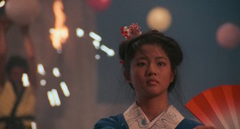 Movie still from “The Karate Kid Part II” (1986), directed by John G. Avildsen – A young woman wearing a blue and white shirt with fireworks in the background; Close Up shot, Low angle