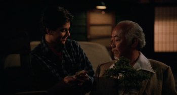 Movie still from “The Karate Kid Part III” (1989), directed by John G. Avildsen – A man sitting next to another man holding a cell phone; Medium shot, Over the shoulder angle