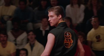 Movie still from “The Karate Kid Part III” (1989), directed by John G. Avildsen – A man in a black vest is standing in front of a group of people; Close Up shot, Low angle