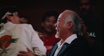 Movie still from “The Karate Kid Part III” (1989), directed by John G. Avildsen – An older man laughing while sitting in front of a group of people; Close Up shot, Over the shoulder angle