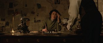 Movie still from “The King's Man” (2021), directed by Matthew Vaughn – A woman sitting at a desk with a pen and paper in front of her face; Medium shot, Over the shoulder angle