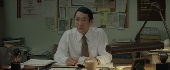 Movie still from “The Kitchen” (2019), directed by Andrea Berloff – A man sitting at a desk in front of papers and a coffee cup; Medium shot, Over the shoulder angle