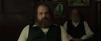 Movie still from “The Kitchen” (2019), directed by Andrea Berloff – A man with a long beard sitting in front of a picture; Medium shot, Over the shoulder angle