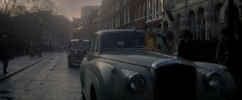 Movie still from “The Last Letter from Your Lover” (2021), directed by Augustine Frizzell – An old car driving down a street past a tall building; Wide shot, Low angle