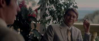 Movie still from “The Last Letter from Your Lover” (2021), directed by Augustine Frizzell – A man wearing a suit and tie sitting in front of some flowers; Close Up shot, Low angle
