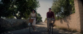Movie still from “The Last Letter from Your Lover” (2021), directed by Augustine Frizzell – A man and a woman riding bikes down a road; Wide shot, High angle