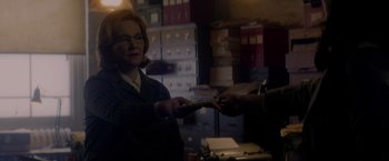 Movie still from “The Last Letter from Your Lover” (2021), directed by Augustine Frizzell – A woman in a dark room handing a card to another person; Medium shot, Over the shoulder angle