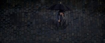 Movie still from “The Last Letter from Your Lover” (2021), directed by Augustine Frizzell – A person sitting on a brick wall holding an umbrella; Extreme Wide shot, Overhead angle
