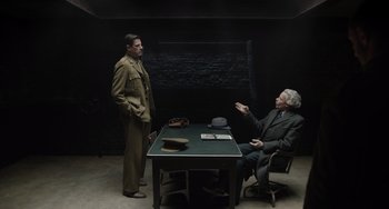 Movie still from “The Last Vermeer” (2019), directed by Dan Friedkin – Two men are sitting at a table in a dark room; Wide shot, Over the shoulder angle