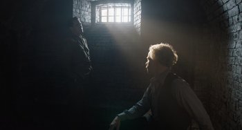 Movie still from “The Last Vermeer” (2019), directed by Dan Friedkin – Two people in a room with a brick wall and a brick window; Medium shot, Low angle