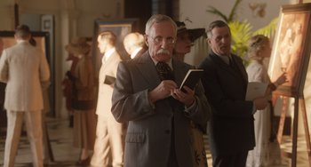 Movie still from “The Last Vermeer” (2019), directed by Dan Friedkin – An older man in a suit and tie holding a book; Medium shot, Low angle
