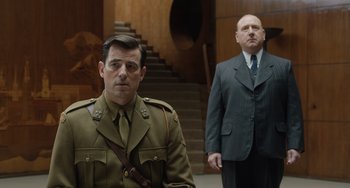 Movie still from “The Last Vermeer” (2019), directed by Dan Friedkin – A man in a military uniform standing next to another man in a business suit; Medium shot, Over the shoulder angle