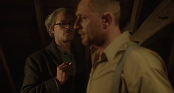 Movie still from “The Last Vermeer” (2019), directed by Dan Friedkin – A man standing next to another man holding a cigarette in his mouth; Medium shot, Over the shoulder angle