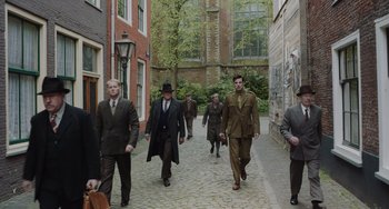 Movie still from “The Last Vermeer” (2019), directed by Dan Friedkin – A group of men walking down a brick street; Wide shot, Over the shoulder angle