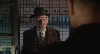 Movie still from “The Last Vermeer” (2019), directed by Dan Friedkin – An older man wearing a suit and a hat is talking to a young man; Medium shot, Over the shoulder angle