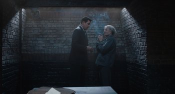 Movie still from “The Last Vermeer” (2019), directed by Dan Friedkin – Two people standing in front of a brick wall and a table; Medium shot, Over the shoulder angle