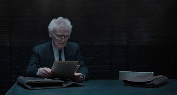 Movie still from “The Last Vermeer” (2019), directed by Dan Friedkin – An older man sitting at a table looking at papers; Medium shot, Over the shoulder angle